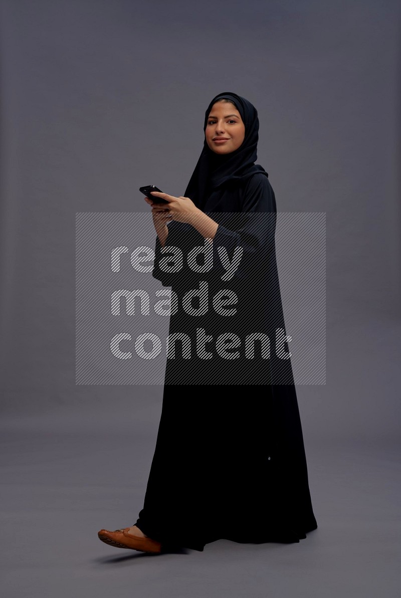 Saudi woman wearing Abaya standing texting on phone on gray background