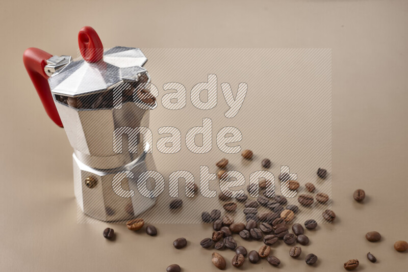 A moka pot with red handle surrounded by roasted coffee beans on beige background
