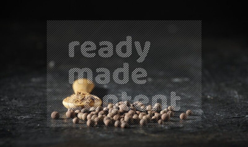 A wooden spoon full of allspice powder and whole balls spreaded on a textured black flooring