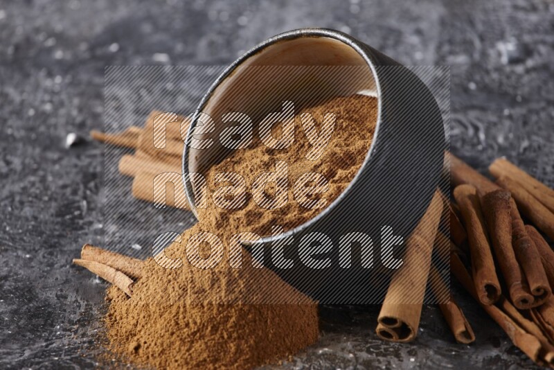 Black pottery bowl over filled with cinnamon powder and cinnamon sticks around the bowl on a textured black background