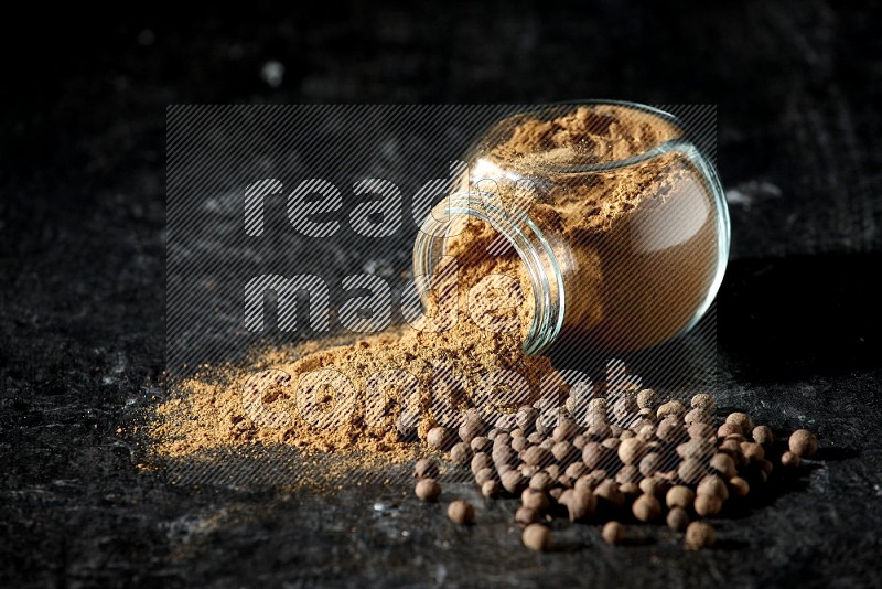 A flipped glass spice jar full of allspice powder and powder spilled out of it with whole balls on a textured black flooring