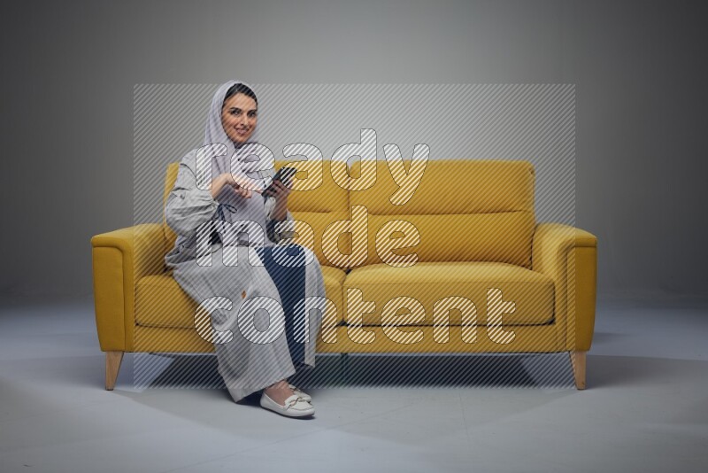 A Saudi woman wearing a light gray Abaya and white head scarf sitting on a yellow sofa and using her phone eye level on a grey background