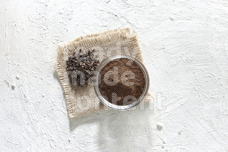 A glass bowl full of cloves powder and cloves grains on a burlap piece on a white flooring