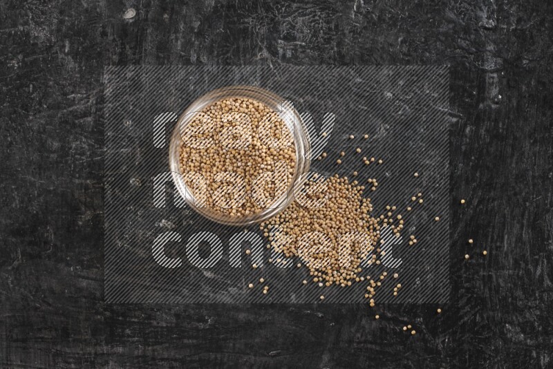 A glass jar full of mustard seeds and jar is flipped and seeds spread out on a textured black flooring
