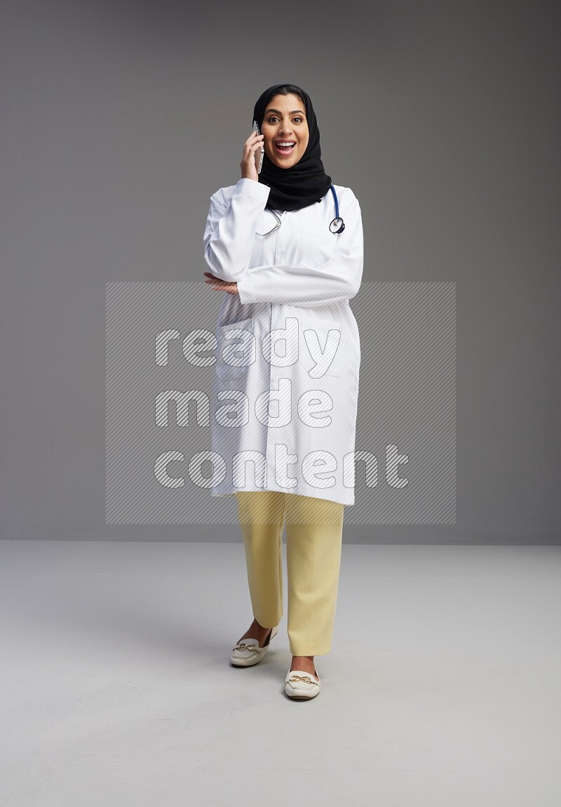 Saudi woman wearing lab coat with stethoscope standing talking on phone on Gray background