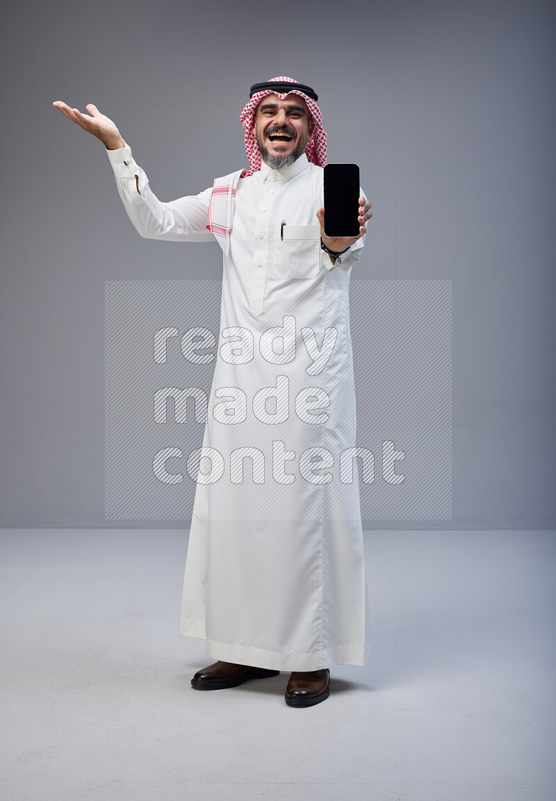 Saudi man Wearing Thob and red Shomag standing showing phone to camera on Gray background