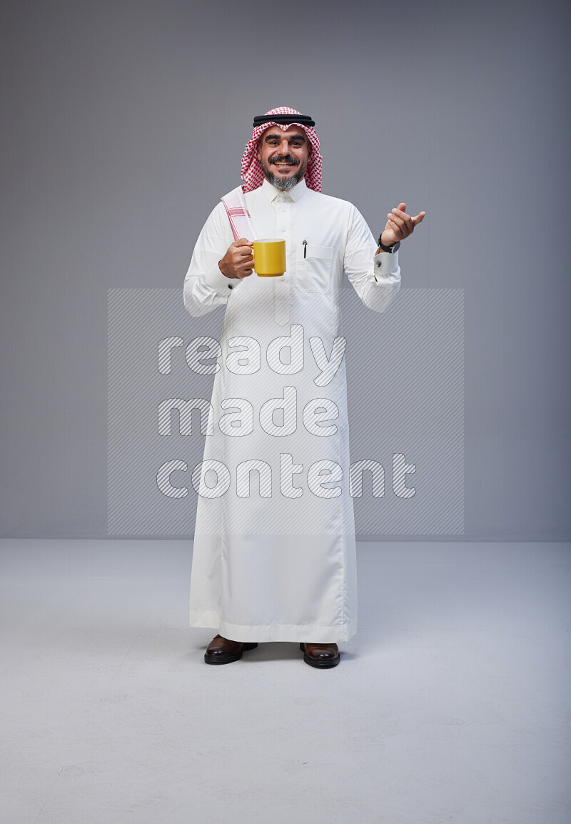 Saudi man Wearing Thob and red Shomag standing holding mug on Gray background