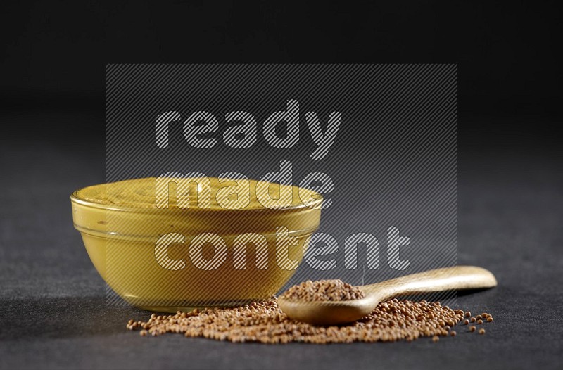 A glass bowl full of mustard paste with mustard seeds underneath and a full wooden spoon on black flooring