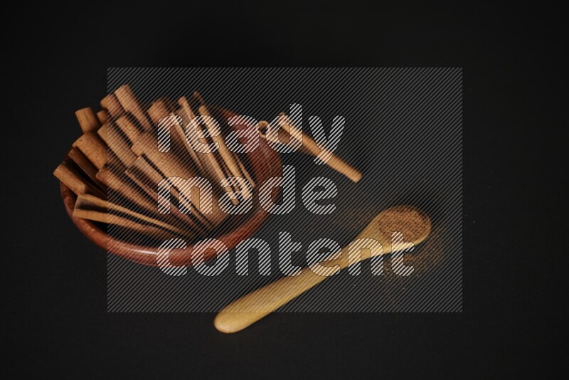 Cinnamon sticks in wooden bowl and cinnamon powder in a wooden spoon on black background