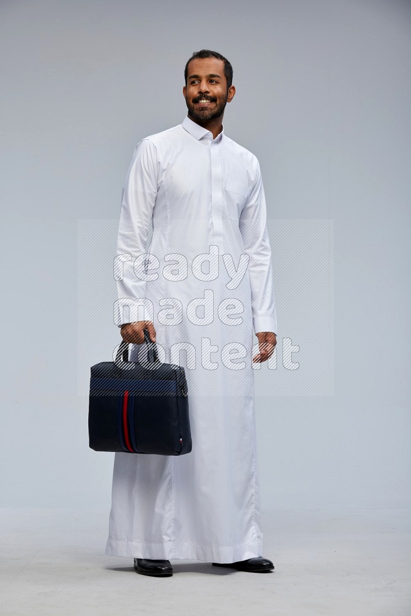 Saudi man wearing thob standing holding suitcase on gray background
