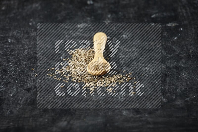 A wooden spoon full of cumin powder and cumin seeds spreaded on textured black flooring