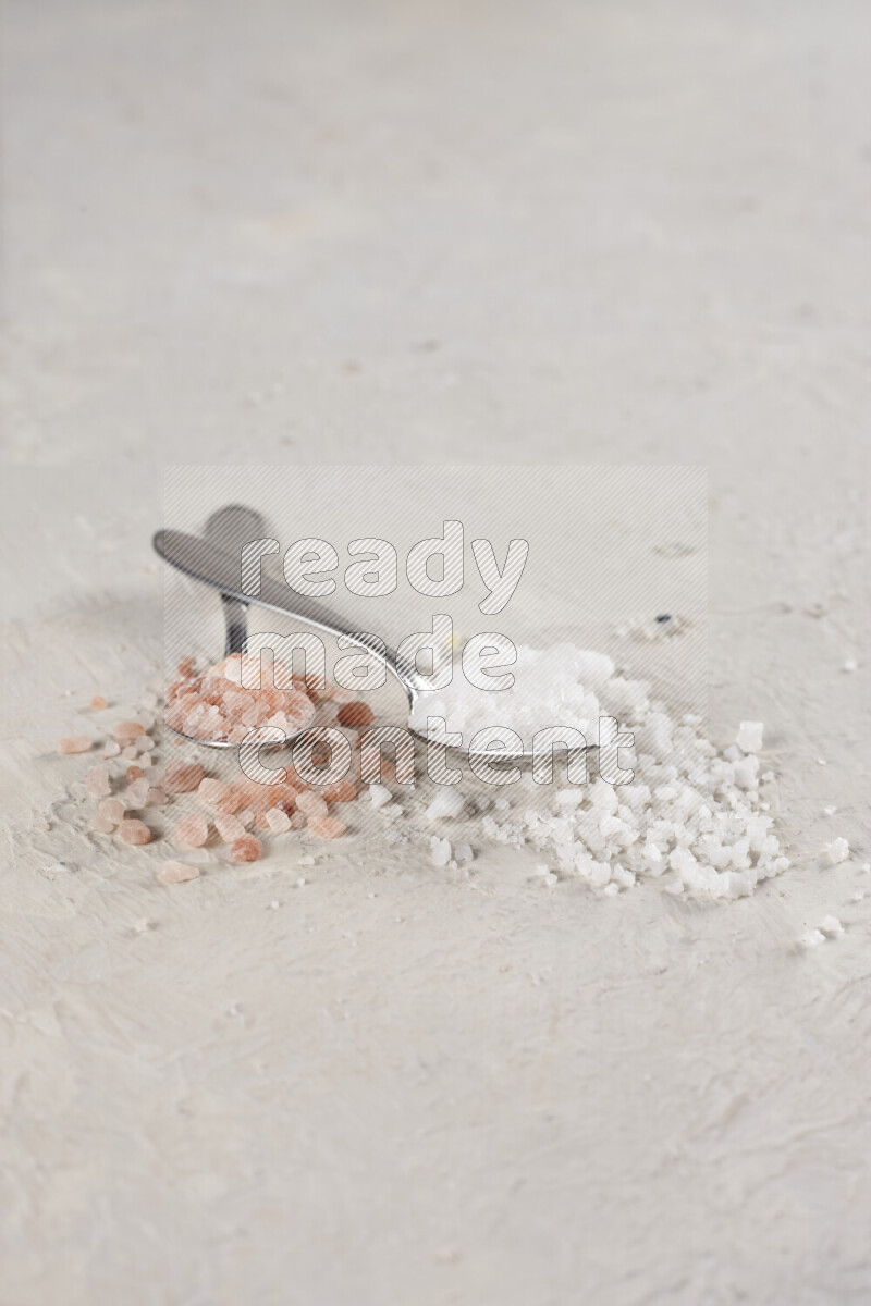 2 metal spoons filled with white salt and pink himalayan salt on white background