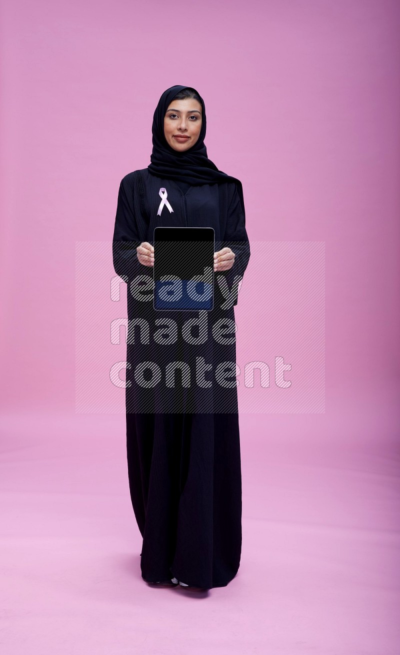 Saudi woman wearing pink ribbon on Abaya standing showing tablet to camera on pink background