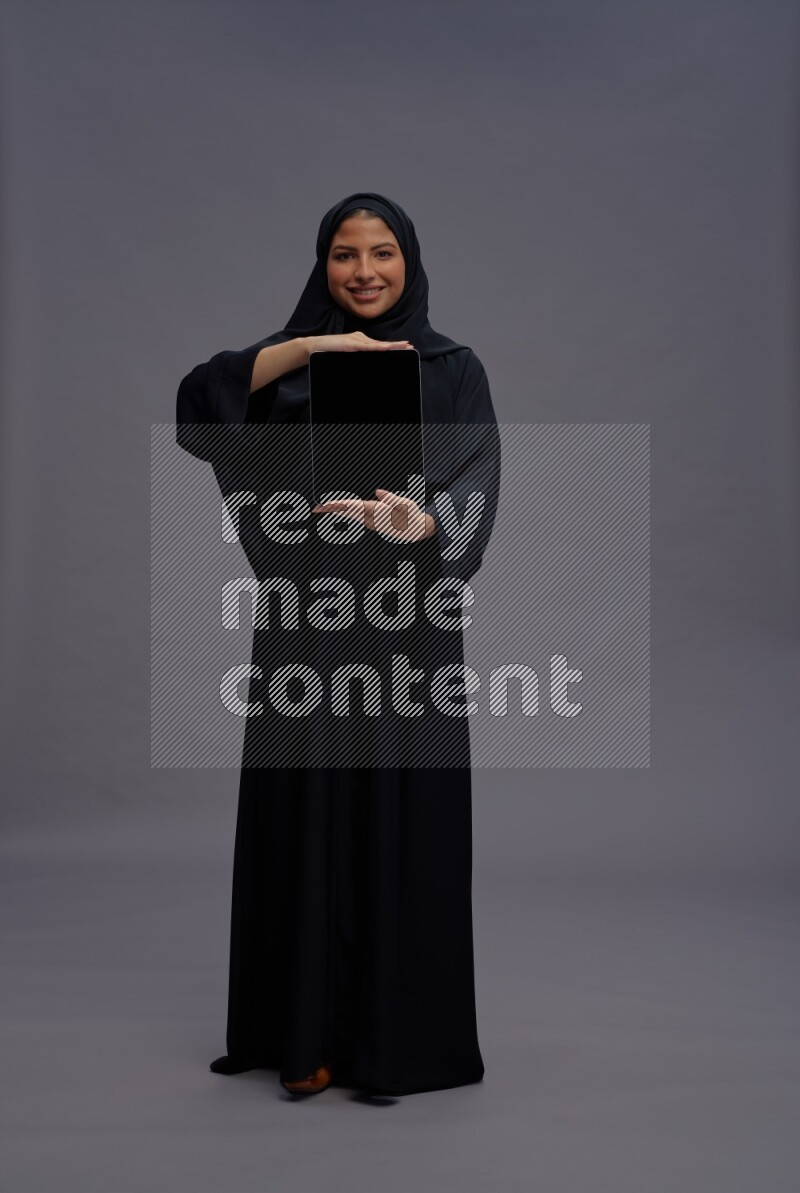 Saudi woman wearing Abaya standing showing tablet to camera on gray background