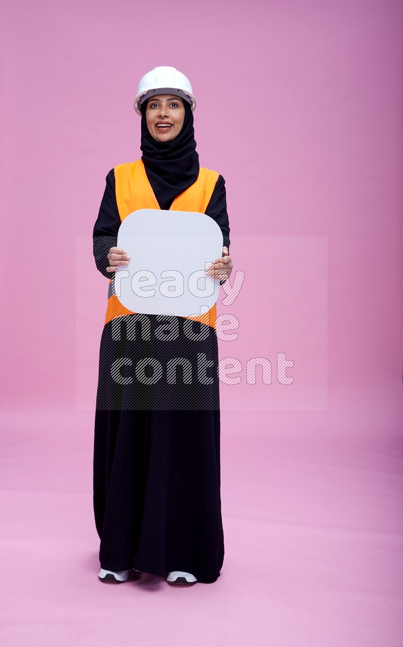 Saudi woman wearing Abaya with engineer vest and helmet standing holding social media sign on pink background
