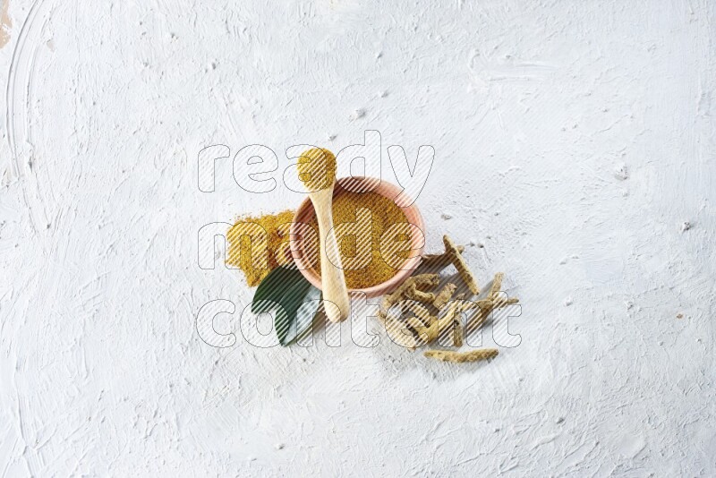 A wooden bowl and wooden spoon full of turmeric powder with dried turmeric fingers beside it on textured white flooring