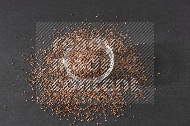 A glass bowl full of flaxseeds surrounded by the seeds on a black flooring