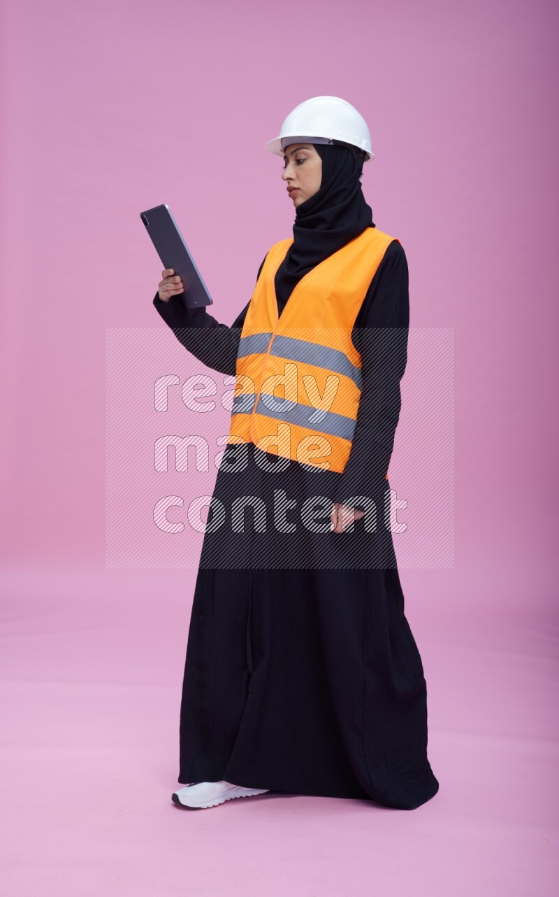 Saudi woman wearing Abaya with engineer vest and helmet standing working on tablet on pink background