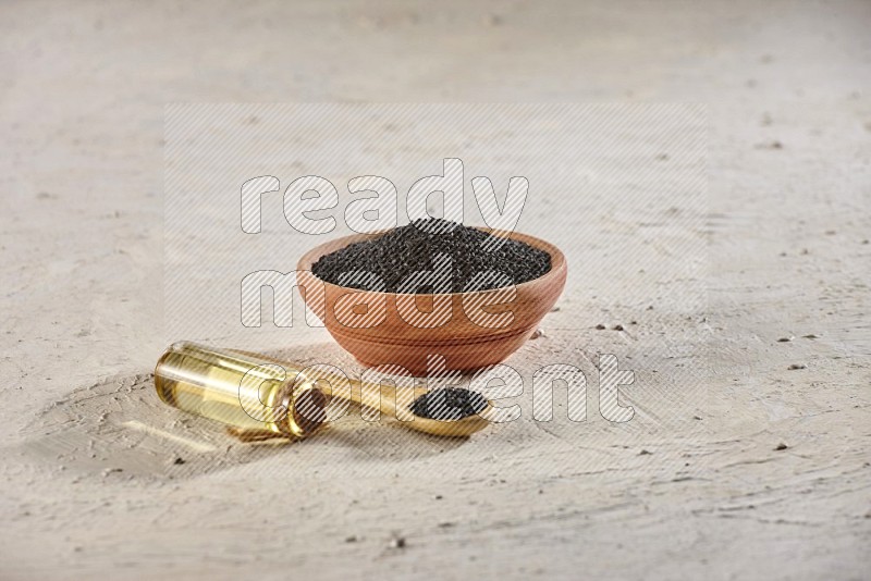 A wooden bowl and spoon full of black seeds with a bottle of black seeds oil on a textured white flooring