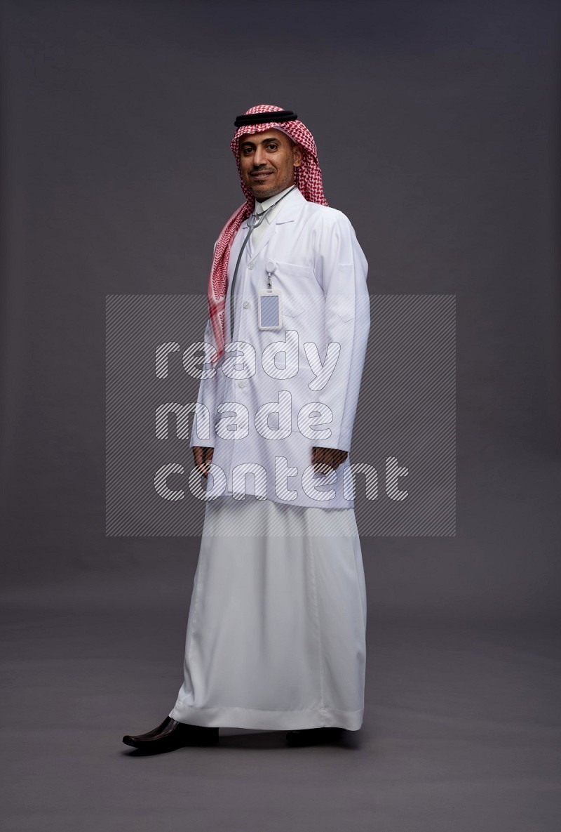Saudi man wearing thob with lab coat and shomag with pocket employee badge with stethoscope standing interacting with the camera on gray background