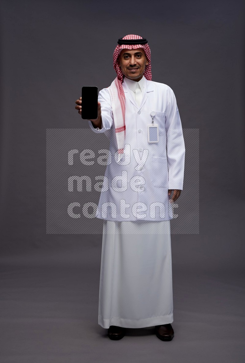 Saudi man wearing thob with lab coat and shomag with pocket employee badge standing showing phone to camera on gray background