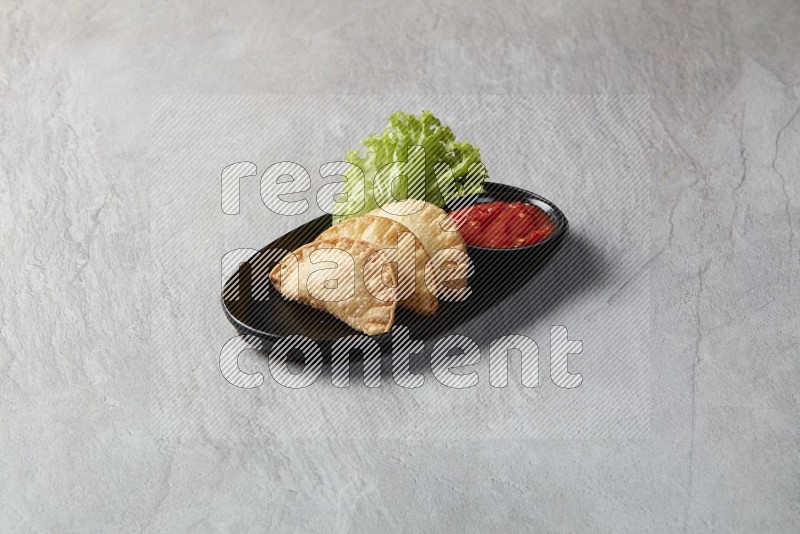 Three fried sambosas in an oval shaped black plate and a red sauce in a black round ramekin on a gray background