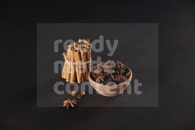A stacked and bounded cinnamon sticks and a wooden bowl full of star anise on a black background