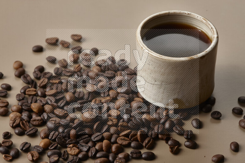 A beige pottery cup of coffee surrounded by roasted coffee beans on beige background