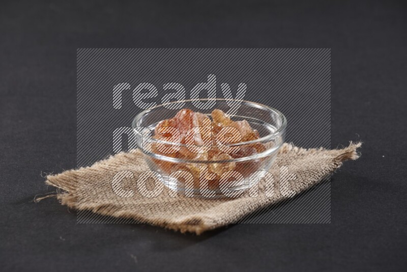 A glass bowl full of gum arabic on a burlap piece on black flooring