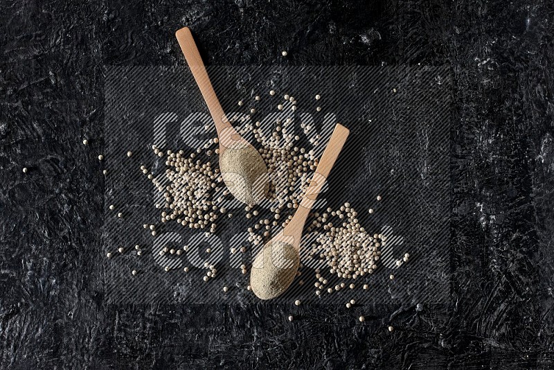 2 wooden spoons full of white pepper powder with white pepper beads on textured black flooring