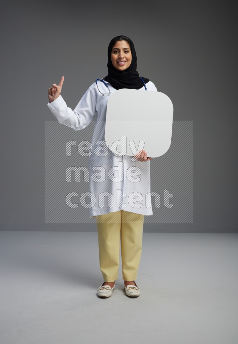 Saudi woman wearing lab coat with stethoscope standing holding social media sign on Gray background