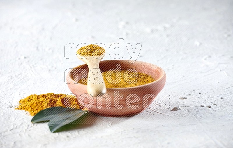 A wooden bowl and wooden spoon full of turmeric powder on textured white flooring