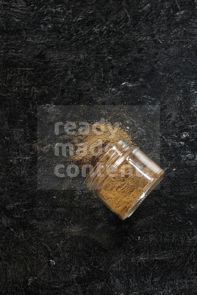 A flipped glass jar full of cumin powder and powder fell out on a textured black flooring