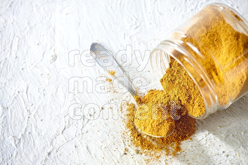 A flipped glass spice jar and a metal spoon full of turmeric powder and powder spilled out of it on textured white flooring