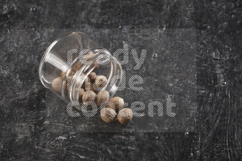 A glass jar full of nutmeg flipped and the seeds spread out on a textured black flooring