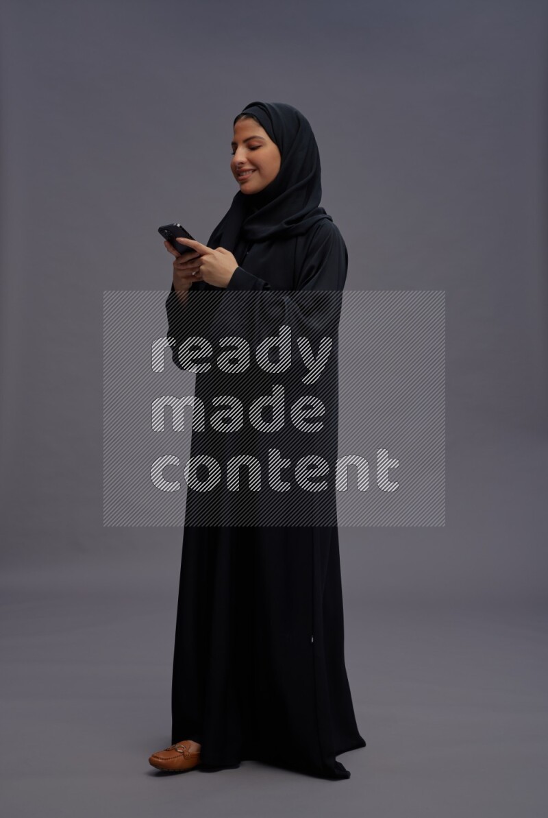 Saudi woman wearing Abaya standing texting on phone on gray background