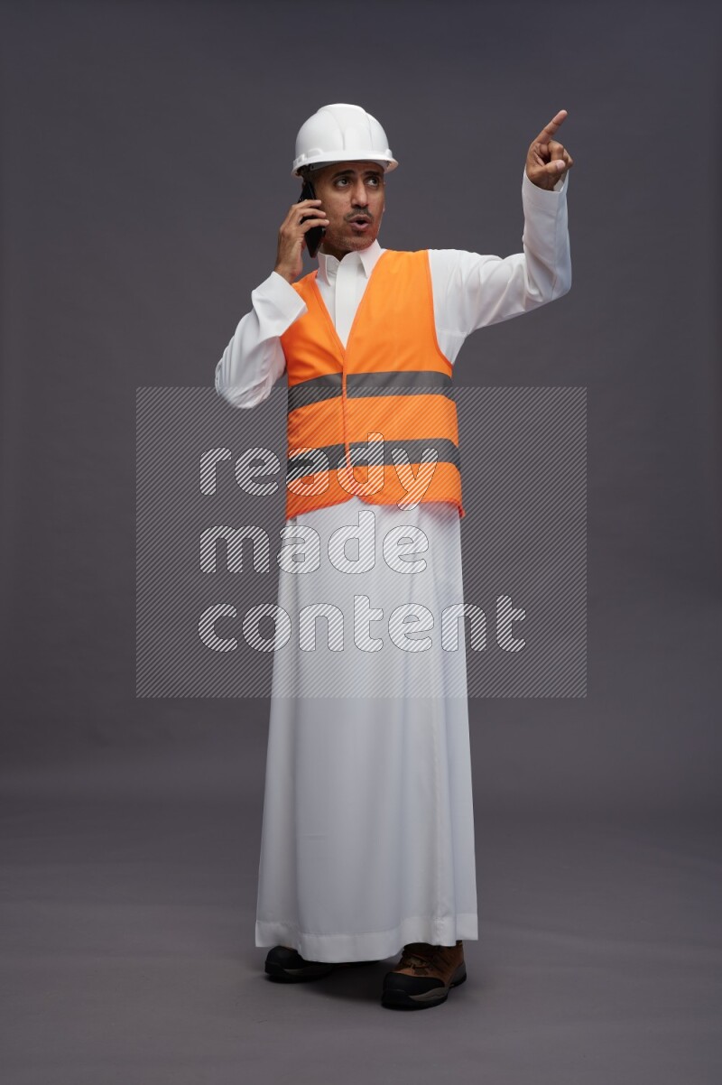 Saudi man wearing thob with engineer vest standing talking on phone on gray background