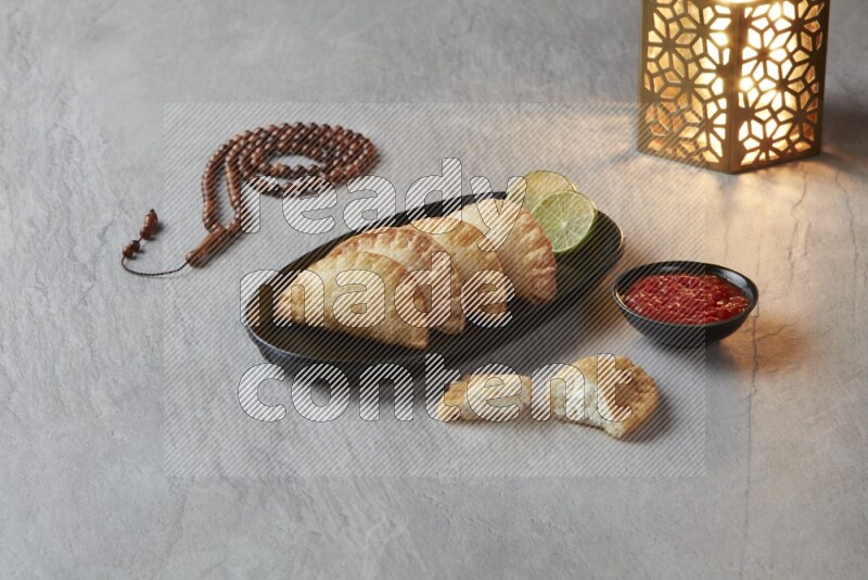 Four fried sambosas in an oval shaped black plate, beside a cut cheese sambosa, a brown misbaha and a golden lantern on a gray background