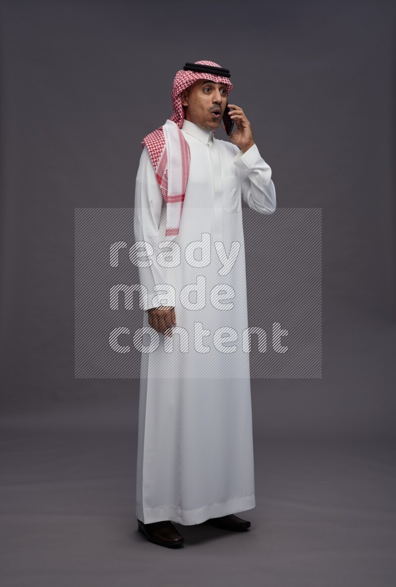 Saudi man wearing thob and shomag standing talking on phone on gray background