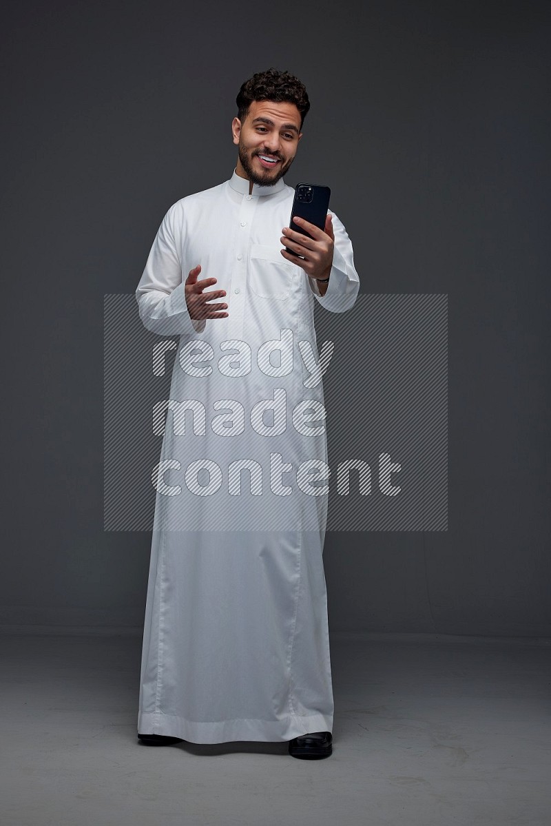 A Saudi man wearing Thobe and making a video call using his phone while standing and making different poses eye level on a gray background