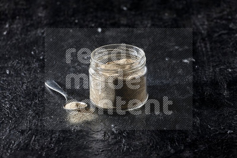 وعاء زجاجي وملعقة معدنية ممتلئان ببودرة الفلفل الأبيض على أرضية سوداء