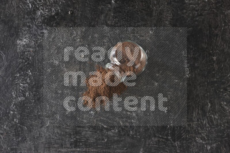 A flipped glass spice jar full of cloves powder and powder came out of it on textured black flooring