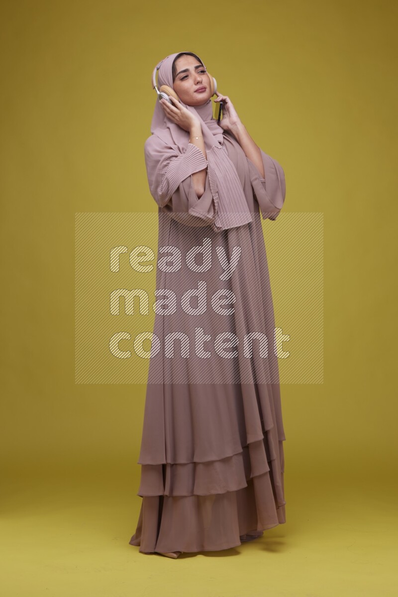 A woman Listening to Music on a Yellow Background wearing Brown Abaya with Hijab