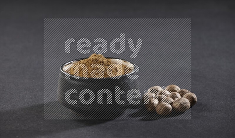 A black pottery bowl full of nutmeg powder with whole seeds beside it on a black flooring