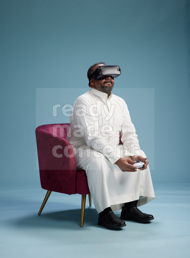 Saudi Man without shimag sitting on chair  gaming with VR and joystick on blue background