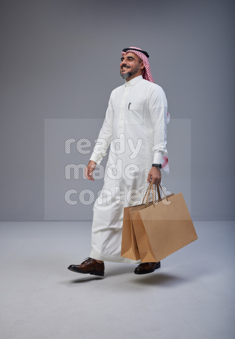 Saudi man Wearing Thob and red Shomag standing holding shopping bag on Gray background