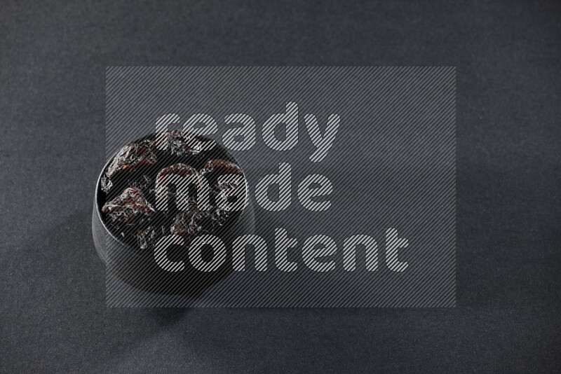 A black pottery bowl full of dried plums on a black background in different angles