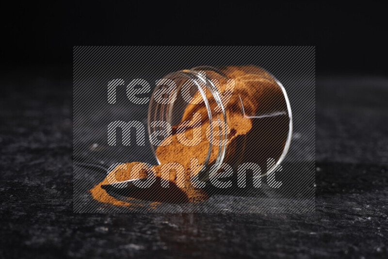 A glass jar full of ground paprika powder flipped with some spilling powder on black background