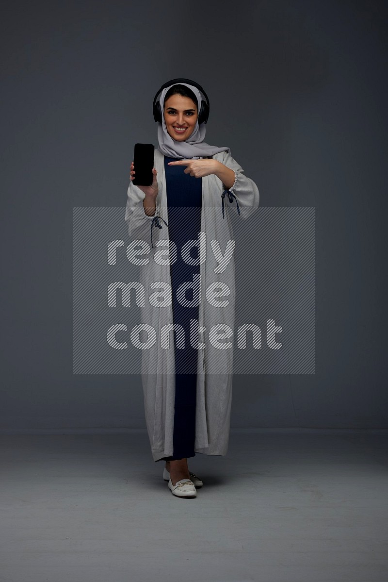 A Saudi woman wearing a light gray Abaya and head scarf setting on a dark grey chair and watching on the tablet eye level on a grey background