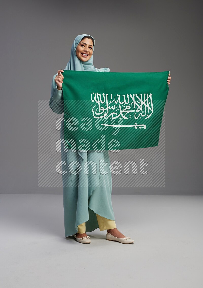 Saudi Woman wearing Abaya standing holding Saudi flag on Gray background